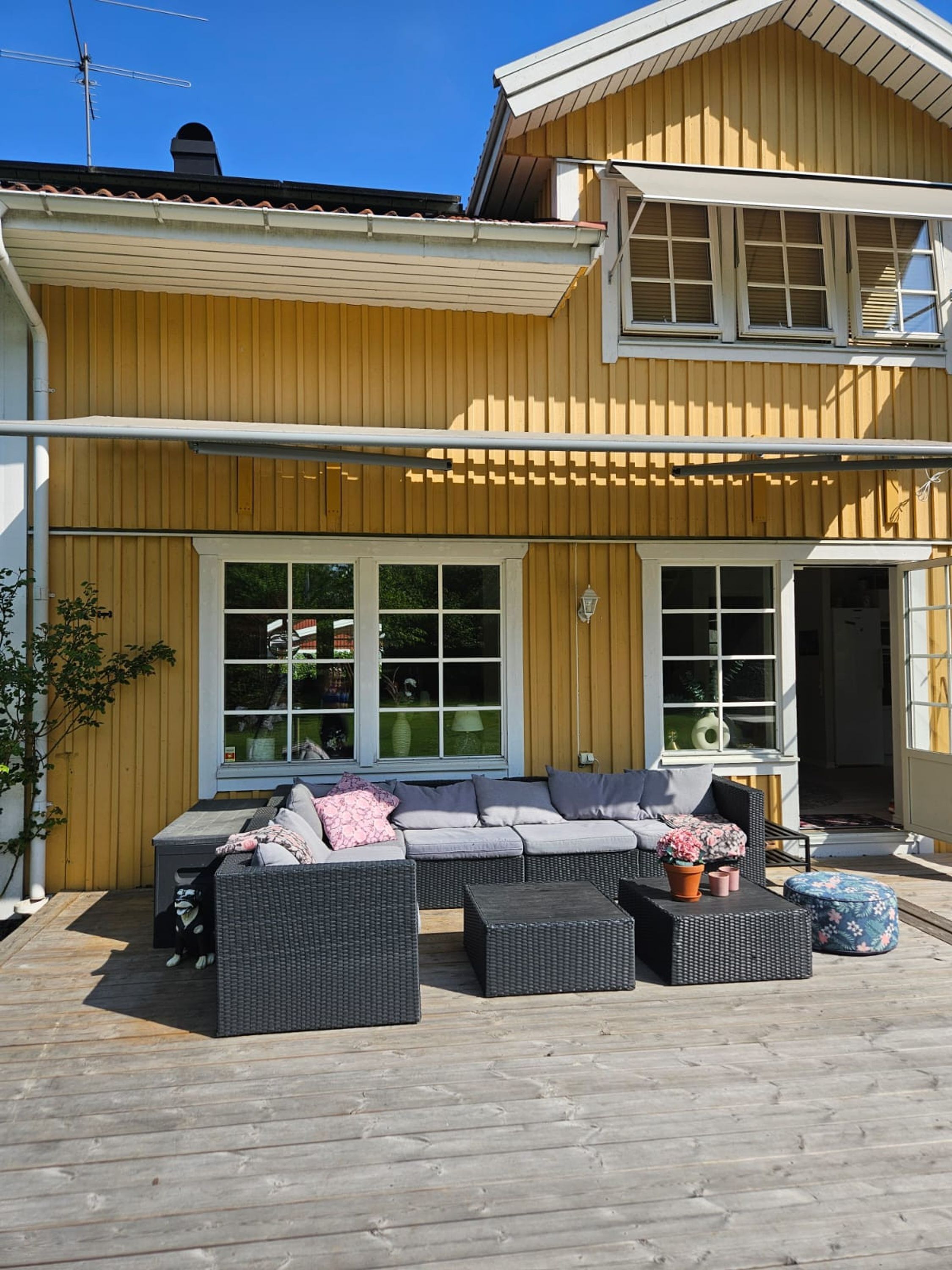 Een geel houten huis met een groot terras, voorzien van rieten meubels en plantenpotten.