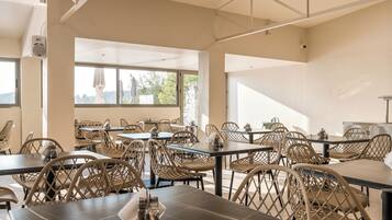 A spacious indoor dining area with high ceilings and large windows, featuring modern wicker chairs and dark tables.