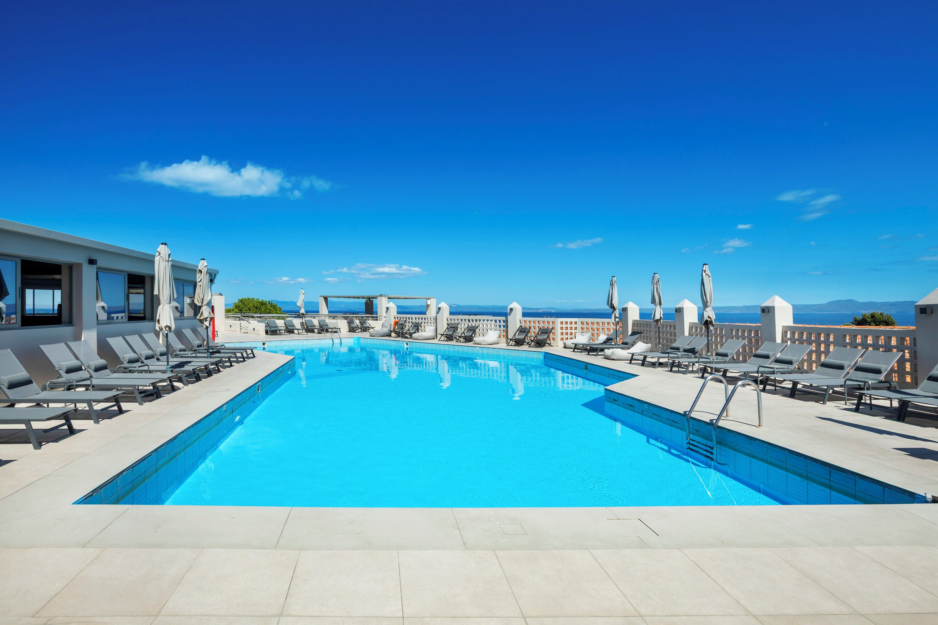 Ein Pool auf dem Dach mit klarem, blauem Wasser, umgeben von Liegestühlen und Sonnenschirmen, unter blauem Himmel.