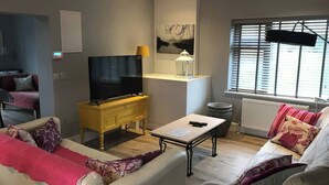 Una sala de estar con un sofá, un televisor sobre un mueble de madera, una mesita con un control remoto y una ventana con persianas.