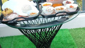 Une table dressée avec une variété de plats, notamment du riz, des plats en sauce et des amuse-gueule, est placée sur une surface de gazon artificiel vert, avec une rambarde métallique en arrière-plan.