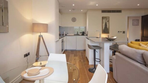 A modern living room with a glass dining table, white chairs, a sofa, and a kitchen area in the background. - Kensington High Street One Bed Flat (London)