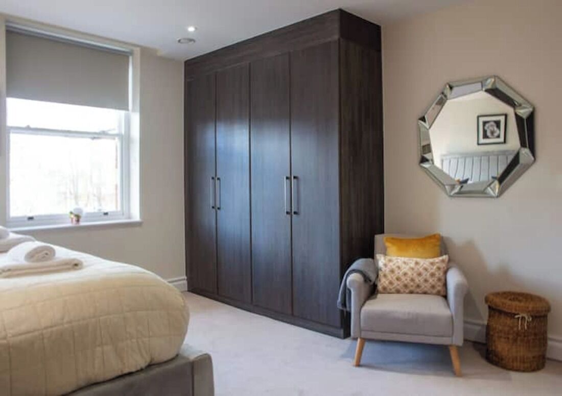 Un dormitorio con un armario grande, una cama con edredón blanco, un sillón gris y un espejo redondo en la pared.