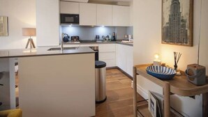 A modern kitchen with white cabinets, a central island, and a dining area with a wooden table and chairs. - Kensington High Street One Bed Flat (London)