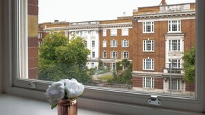 A window with a view of a brick building with multiple windows and a tree in the foreground. - Kensington High Street One Bed Flat (London)