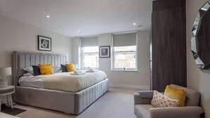 A bedroom with a large bed, a bedside table, a chair, and a mirror on the wall. - Kensington High Street One Bed Flat (London)