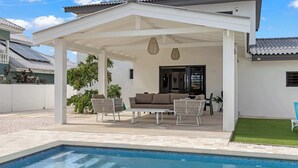 A covered outdoor seating area with a pool, white walls, and a tiled roof.