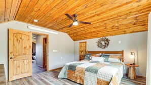 Un dormitorio con techo de madera, una cama con colcha, una mesita de noche y una lámpara fijada a la pared.
