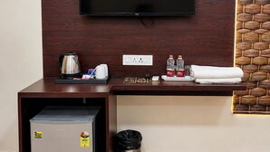 A hotel room with a TV mounted on the wall, a desk with a kettle, coffee maker, and toiletries, a mini-fridge, and a trash bin. - The Manthan Stays (Vrindavan)
