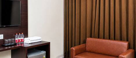 A hotel room with a TV, a desk with a trash can, a brown leather chair, a wooden table, and a curtain.