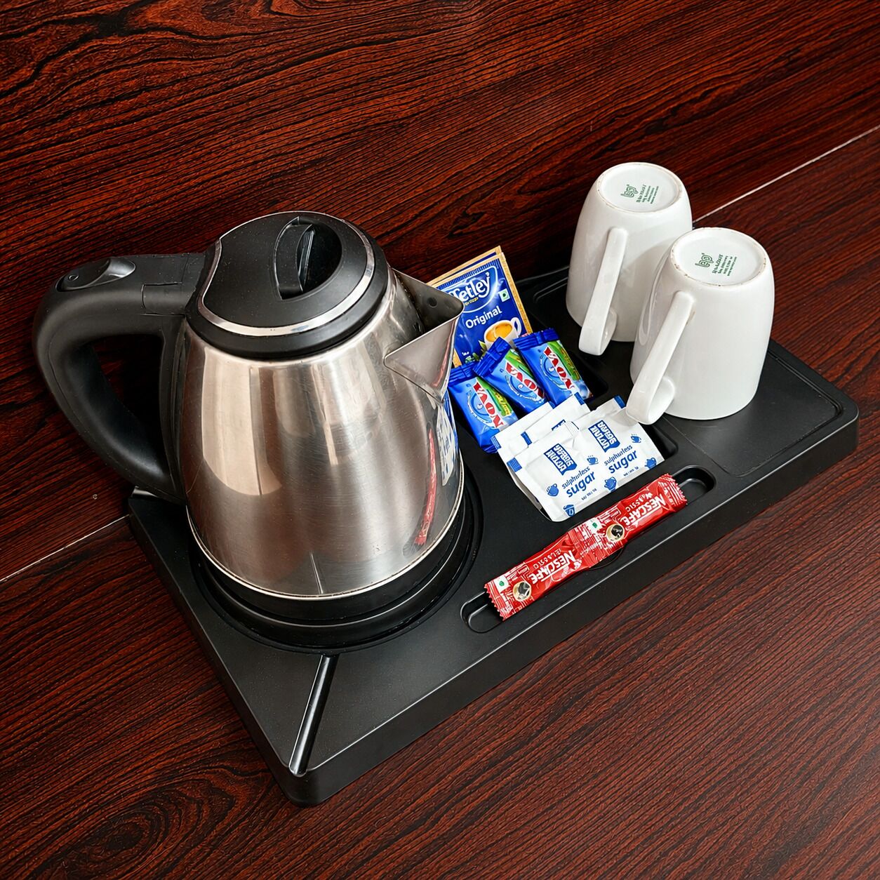 A stainless steel kettle, two white mugs, and packets of instant coffee and sugar on a black tray.