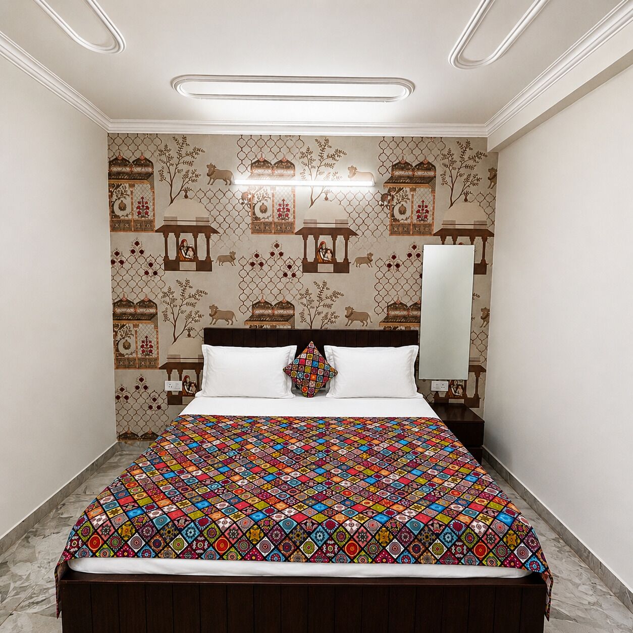 A bedroom with a patterned bedspread, a nightstand with a mirror, and a wall with decorative motifs.