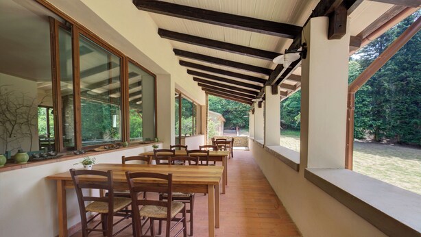 A covered patio with wooden tables and chairs, large windows, and a view of the garden. - Albergo Villa San Michele  (Greve in Chianti)