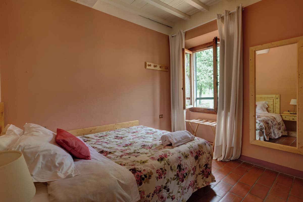 A bedroom with a bed, a bedside table, a mirror, and a window with curtains.
