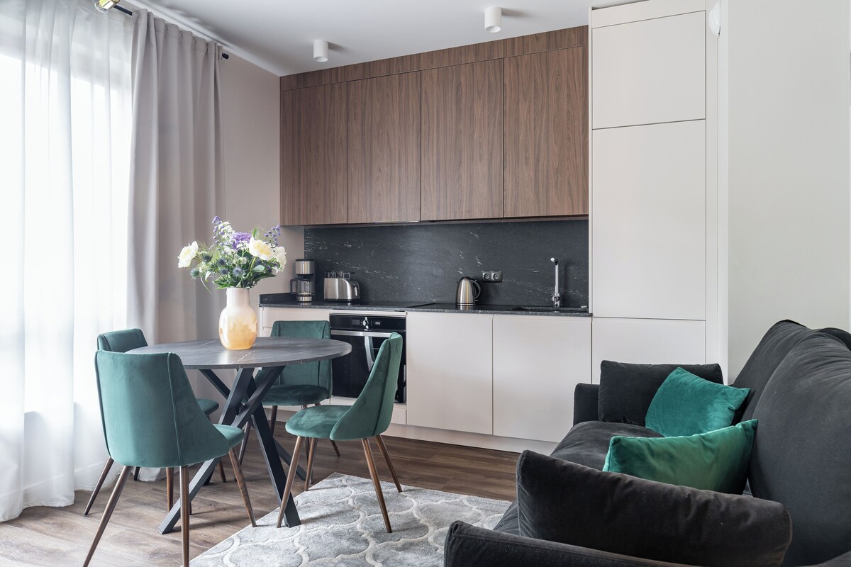 Una cocina moderna con armarios de madera oscura, encimera negra y un rincón comedor con mesa redonda y sillas azul verdosas.