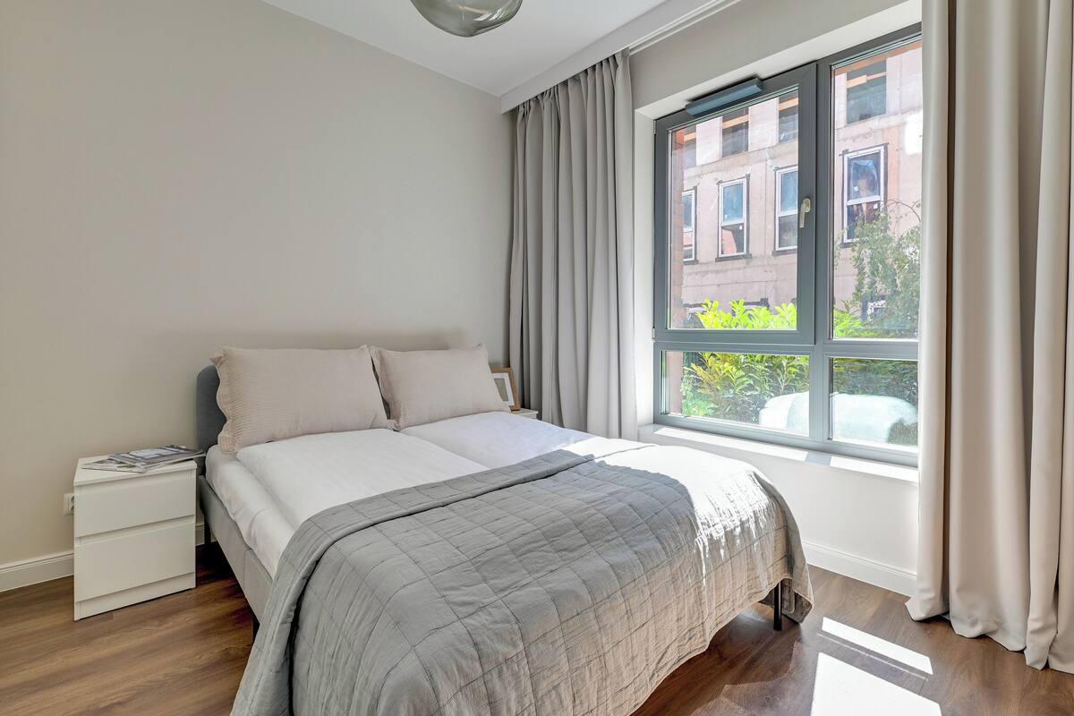 A bedroom with a bed, a nightstand, a window with curtains, and a view of buildings outside.
