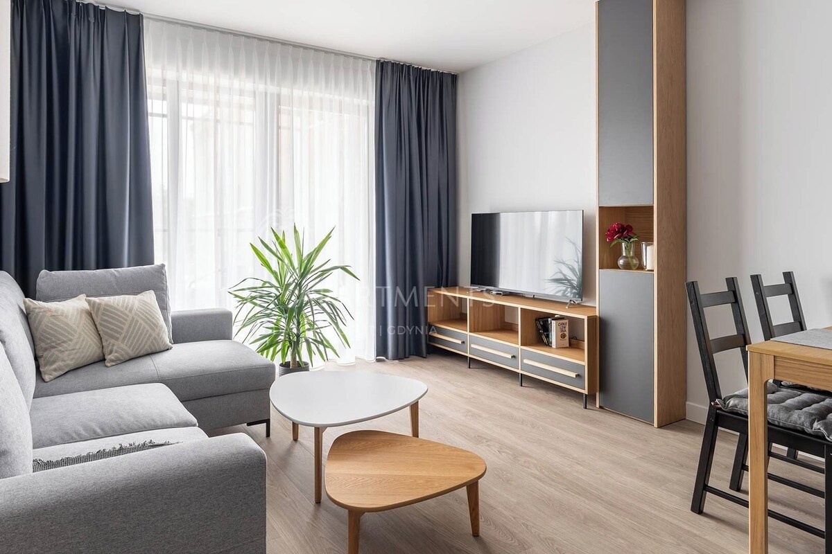 Una sala moderna con un sofá gris, una pequeña mesa redonda, un televisor sobre un mueble de madera, y un comedor con una mesa y sillas de madera.
