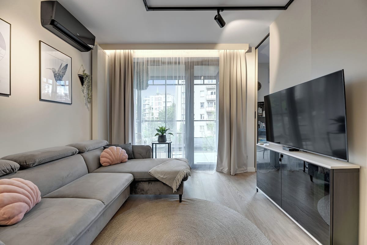 A modern living room with a grey sofa, a flat-screen TV on a cabinet, a large window with sheer curtains, and a small side table with a plant.