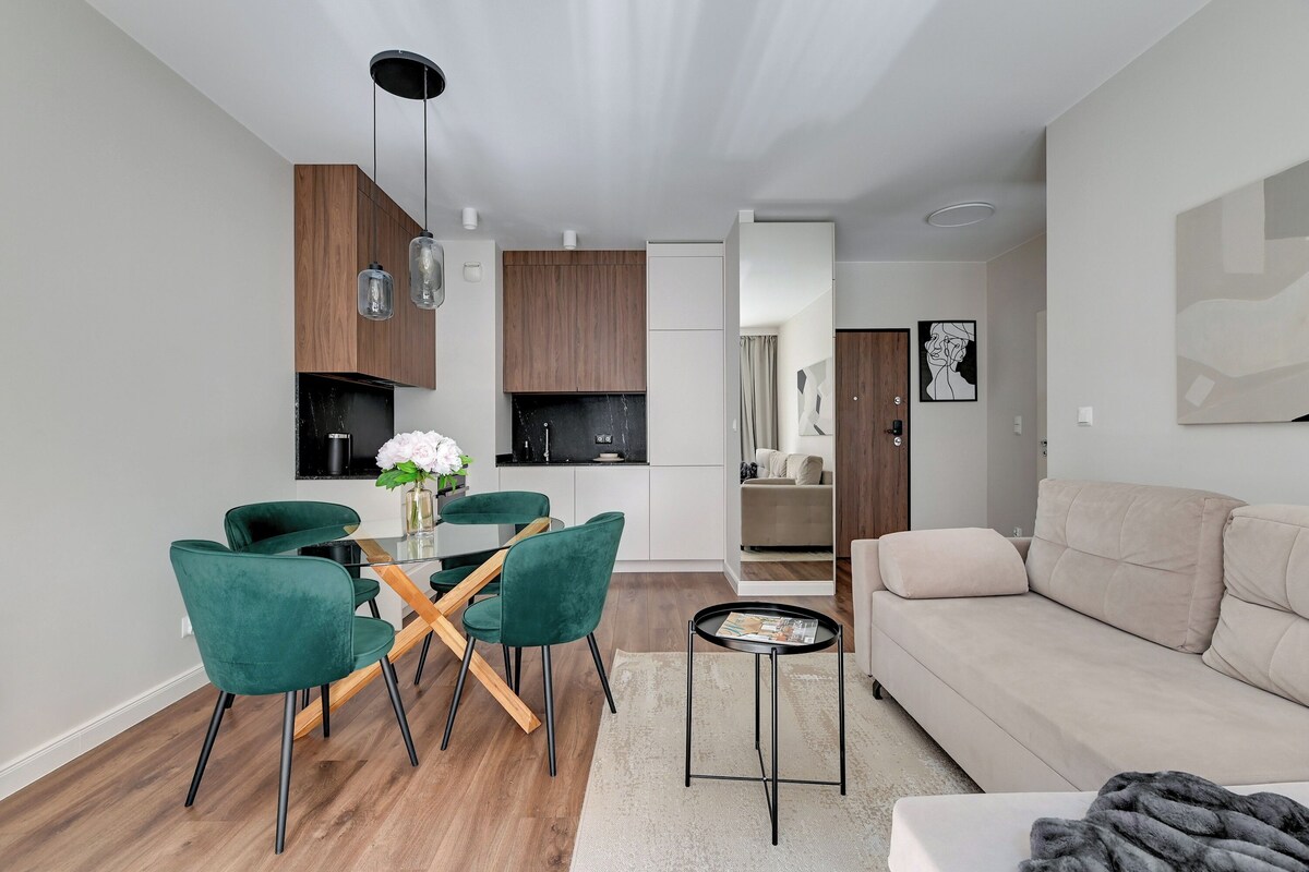 Una sala de estar moderna con un sofá color beige, un comedor con sillas verdes y una cocina con gabinetes de madera y encimera negra.