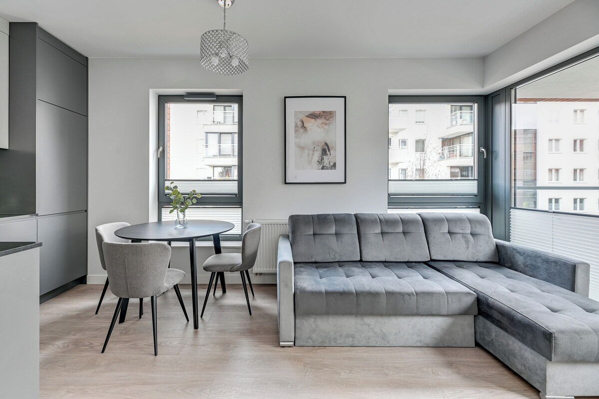 Una sala de estar moderna con un sofá seccional gris, una mesa de comedor con sillas y ventanas amplias con vistas a edificios.