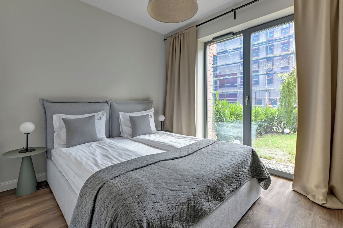 A bedroom with a large bed, two bedside tables, a window with curtains, and a view of a building and greenery outside.