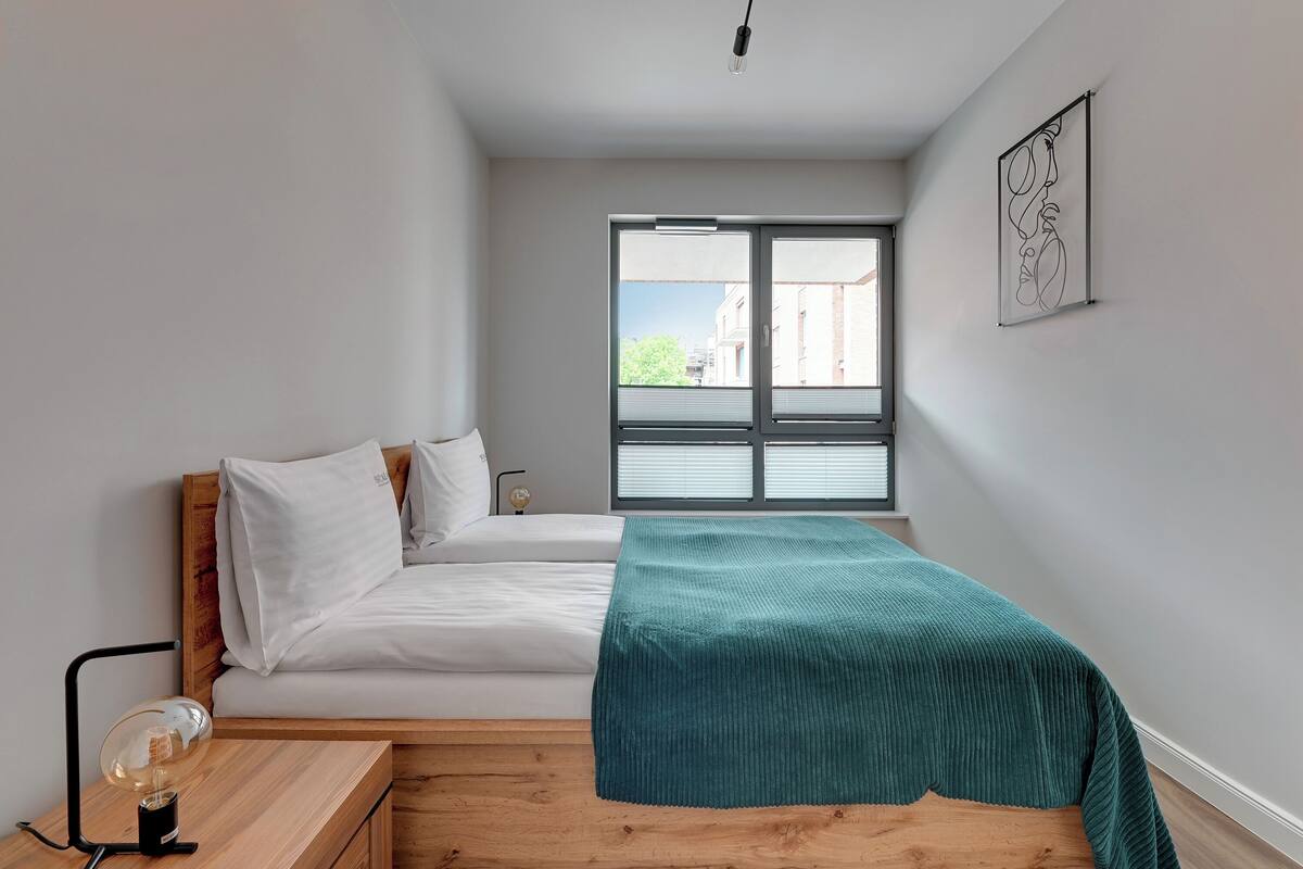 A bedroom with a bed, a bedside table with a lamp, a window with blinds, and a framed artwork on the wall.