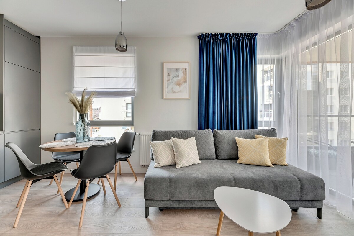 Una sala de estar moderna con un sofá gris, una mesa de centro blanca y sillas negras. Se observa una mesa de comedor con sillas y un jarrón con plantas secas. Se ve una ventana grande con cortinas azules.