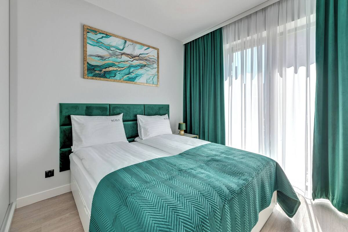 A neatly made bed with a green headboard and white bedding, a painting above the bed, and a window with green curtains.