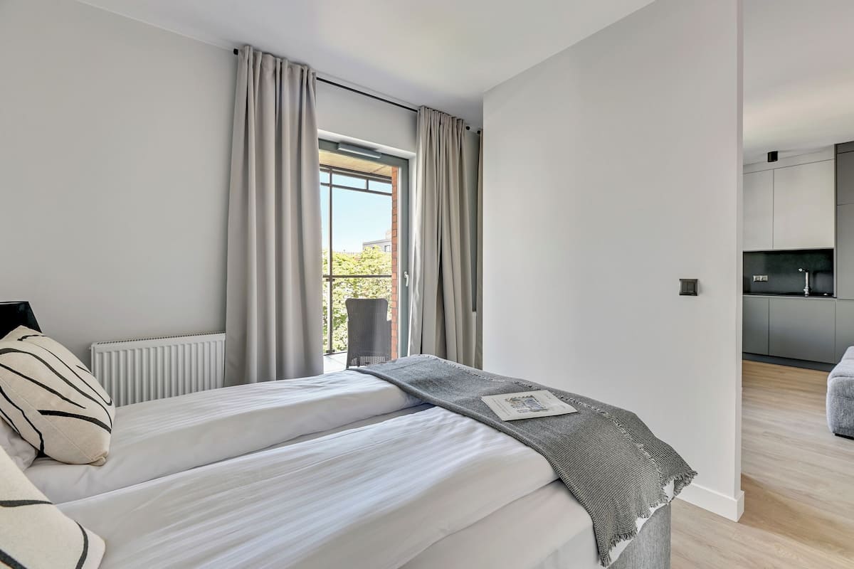 A modern bedroom with a large bed, a view of the outdoors, and a kitchenette in the adjacent room.