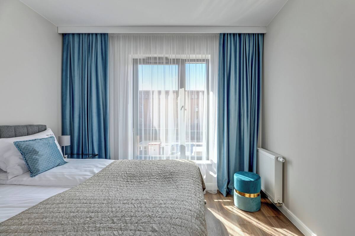 A bedroom with a bed, a bedside table, a lamp, a window with curtains, and a radiator.