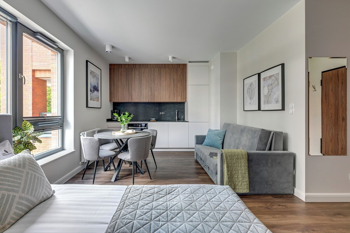 Un espacio habitable moderno y funcional con cama, sofá gris, zona de comedor con mesa y sillas, y kitchenette.