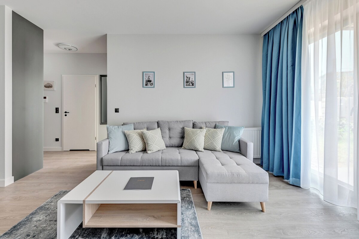 Una sala de estar moderna con un sofá seccional gris, una mesa de centro blanca y un ventanal con cortinas azules.
