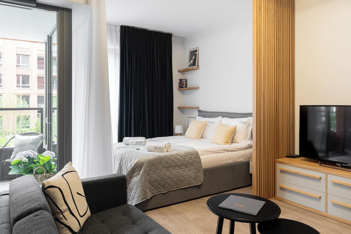 A modern hotel room with a bed, a TV, a sofa, and a window with a view of buildings.