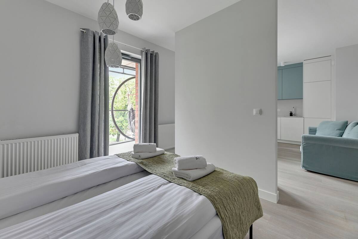A modern hotel room with two beds, a view of the outdoors, and a kitchenette in the background.