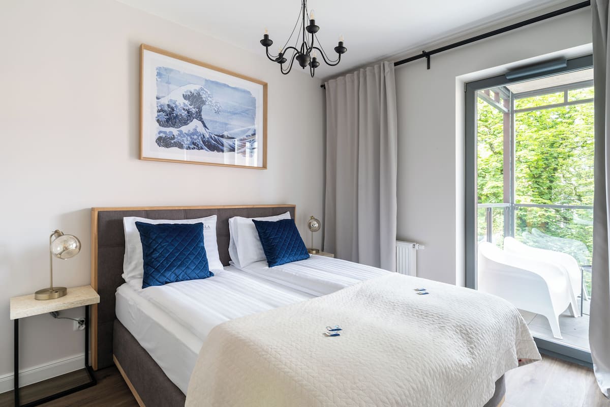 A modern bedroom with a large bed, a bedside table with a lamp, a framed picture on the wall, and a view of greenery through the window.