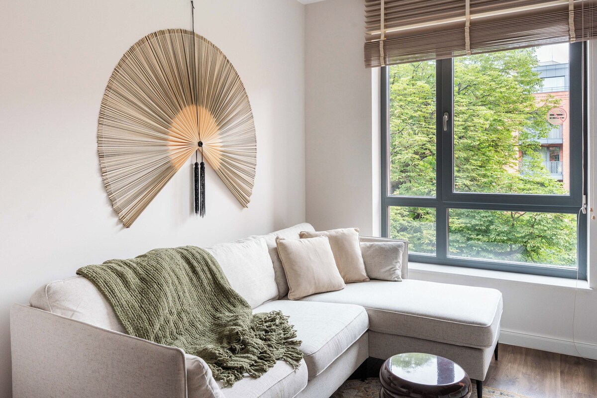 A modern living room with a light-colored sofa, a decorative wall hanging, and a large window with a view of greenery.
