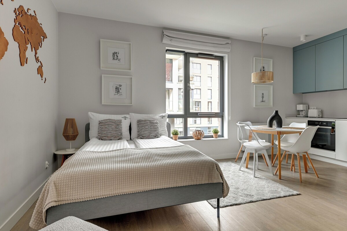 Un dormitorio moderno con cama, mesitas de noche, una ventana con vistas y una zona de kitchenette con mesa y sillas.