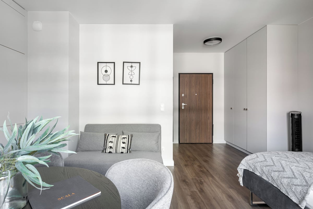 A modern bedroom with a grey sofa, a bed with a patterned blanket, a small table with a book, a potted plant, and two framed abstract artworks on the wall.