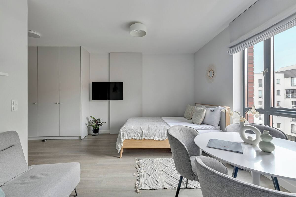 A modern bedroom with a bed, a TV mounted on the wall, a dining table with chairs, and a large window offering a city view.