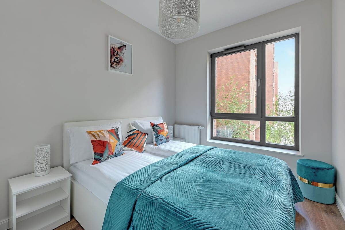 A modern bedroom with a large window, a bed with teal bedding, and a small teal ottoman.