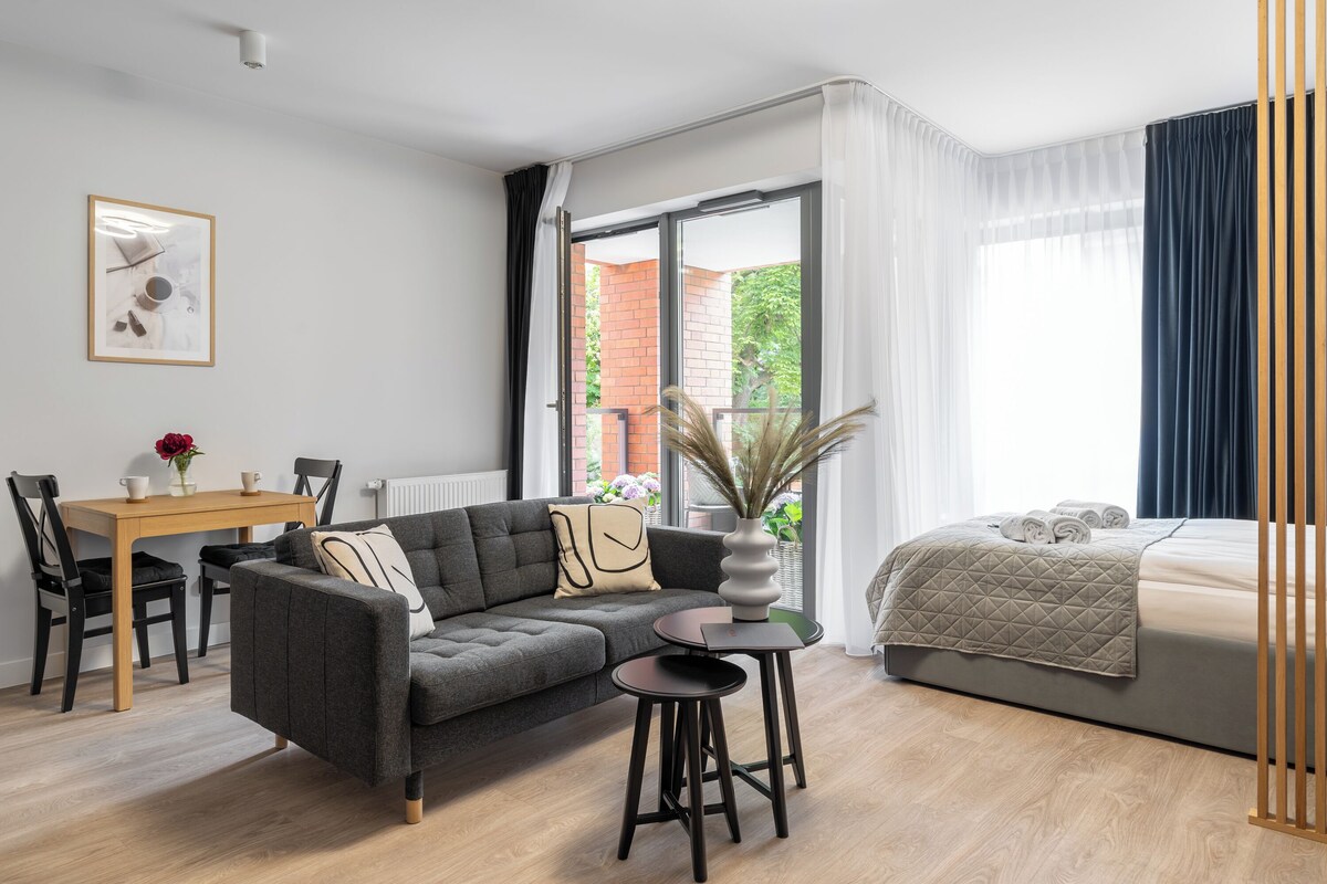 Una habitación moderna y bien iluminada con un sofá gris, una pequeña mesa redonda, una cama con cubrecama gris y vistas a un edificio de ladrillo a través de amplios ventanales.