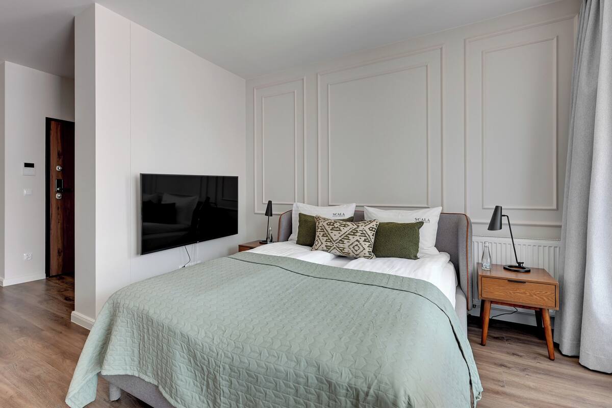 A modern bedroom with a large bed, a TV mounted on the wall, a bedside table with a lamp, and a wooden floor.