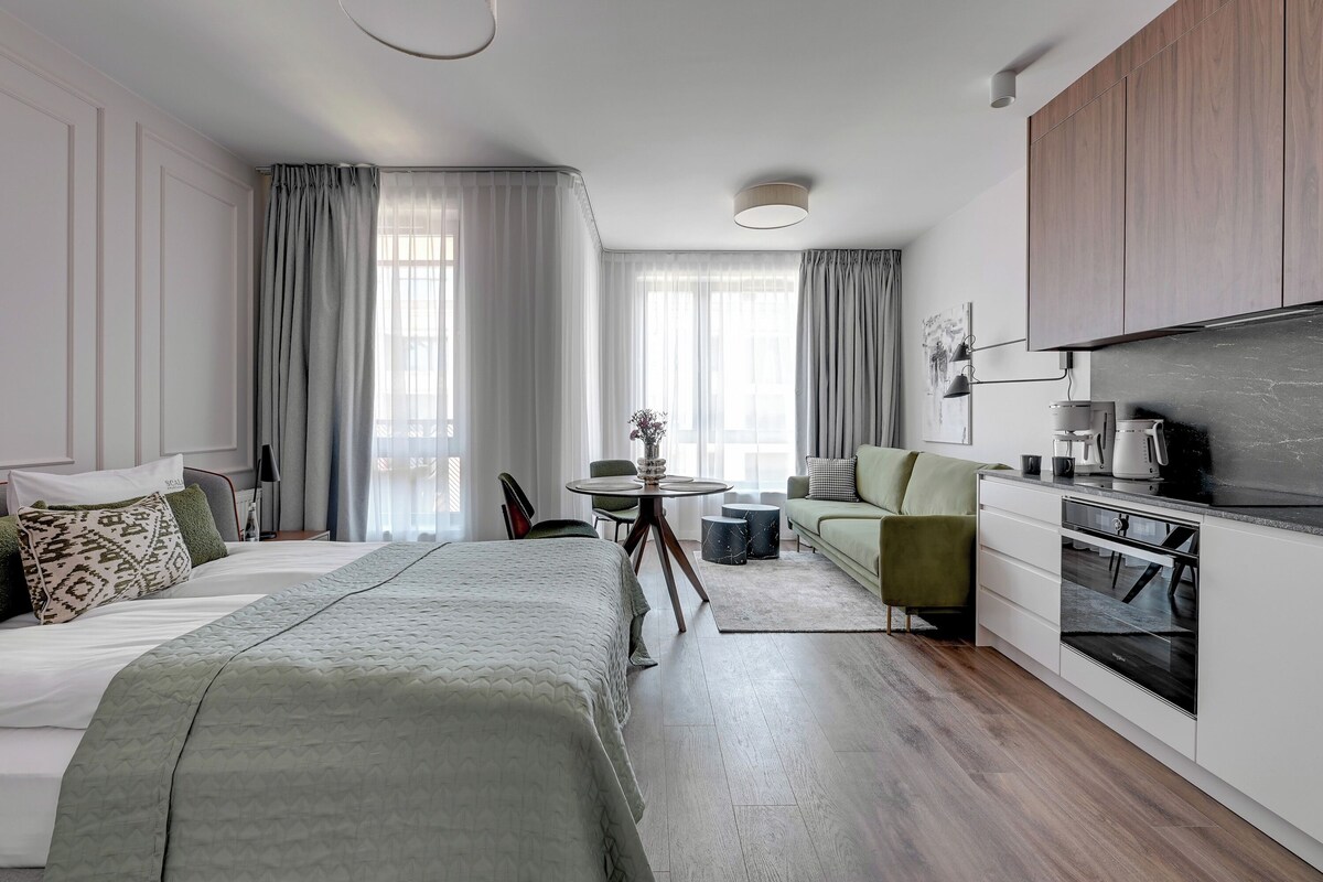Habitación moderna y bien iluminada con cama, sillón verde, una pequeña mesa redonda y un rincón con kitchenette.