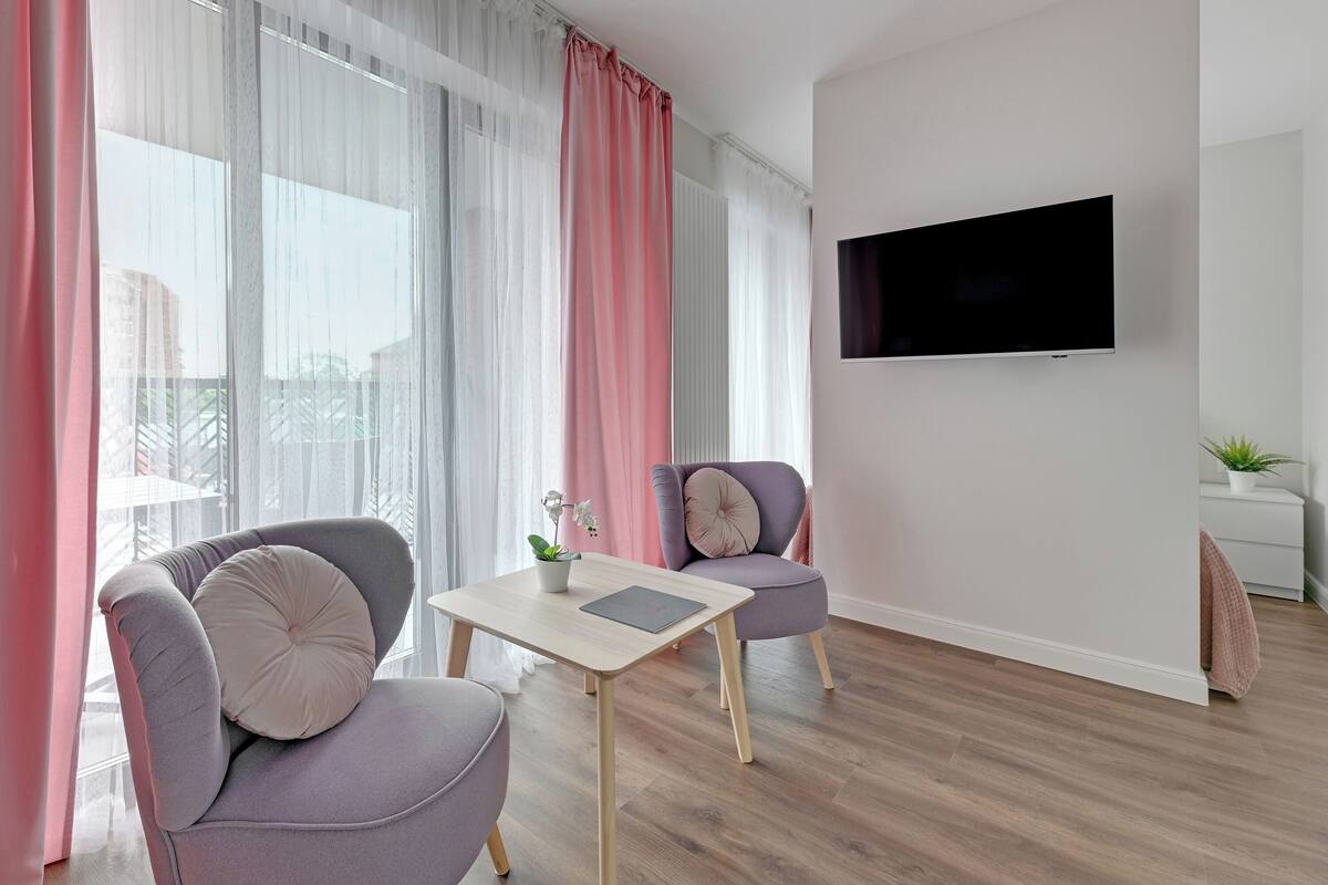 A modern living room with a flat-screen TV mounted on the wall, two grey armchairs with cushions, a small table with a potted plant, and sheer curtains.