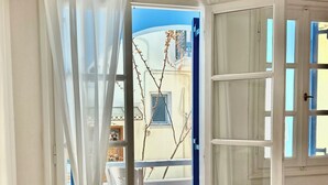 Een slaapkamer met een bed, een nachtkastje en een stoel. Er is een raam met witte gordijnen, en een uitzicht op het buitenbalkon.