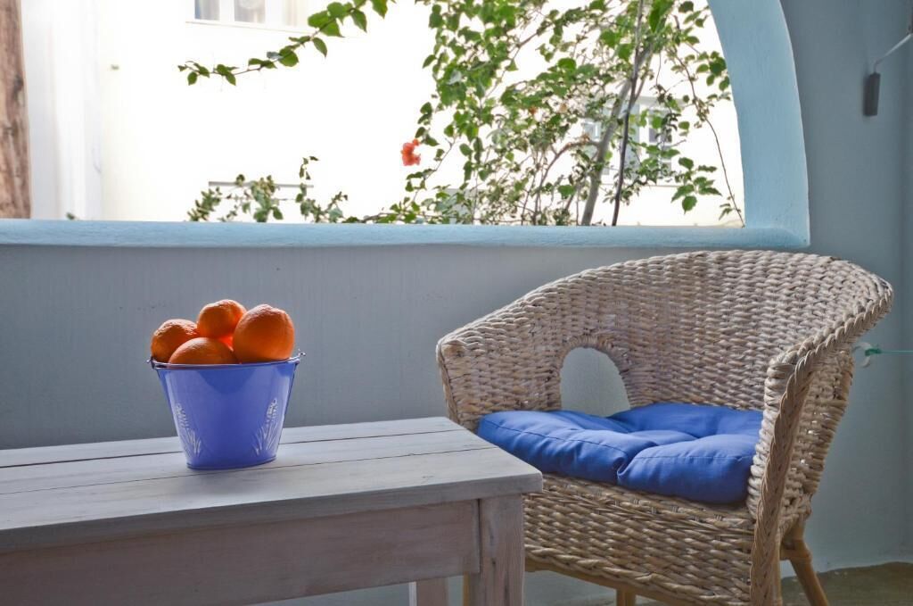 A wicker chair with a blue cushion by a window with a view of greenery and a blue bucket with oranges on a wooden table.