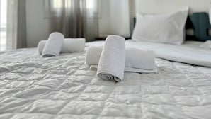 Een net opgemaakt bed met witte lakens en opgerolde handdoeken, een raam met gordijnen, en een witte muur met lichtschakelaar en een klein fotolijstje.
