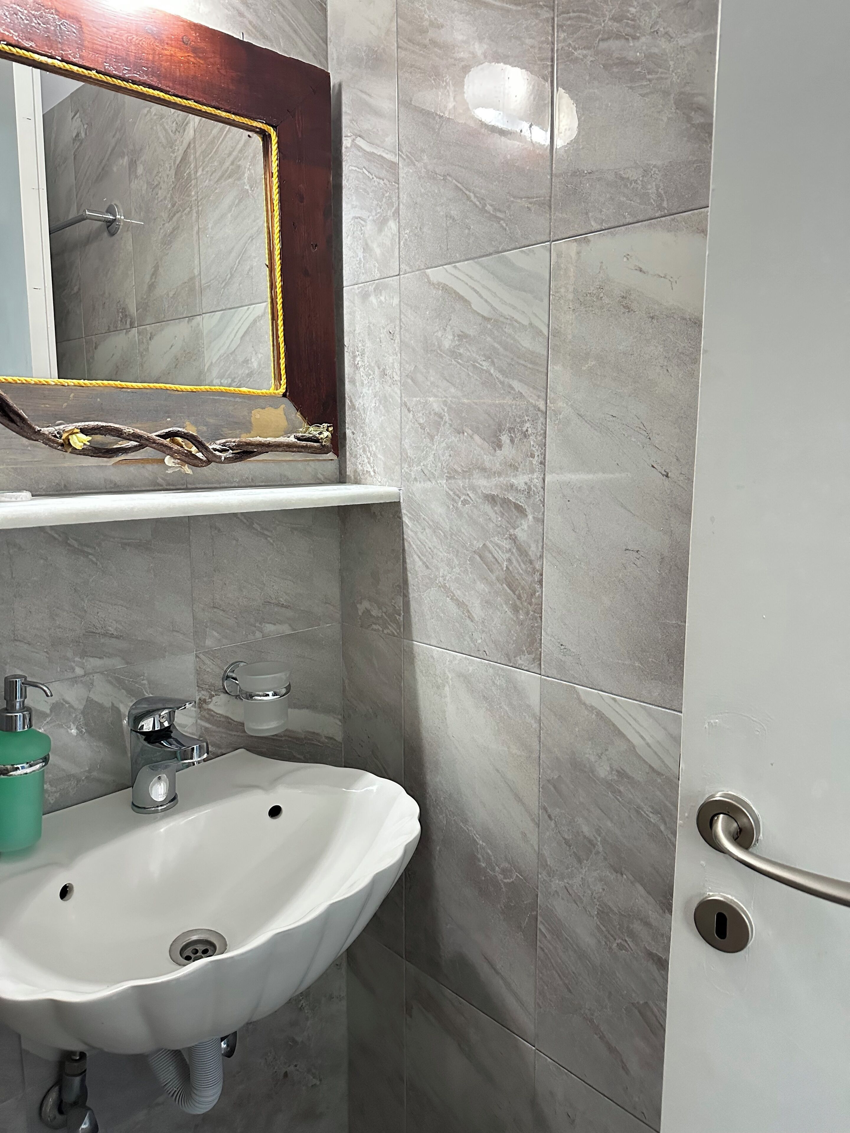Un baño con lavamanos blanco, espejo con marco de madera y baldosas de mármol gris.