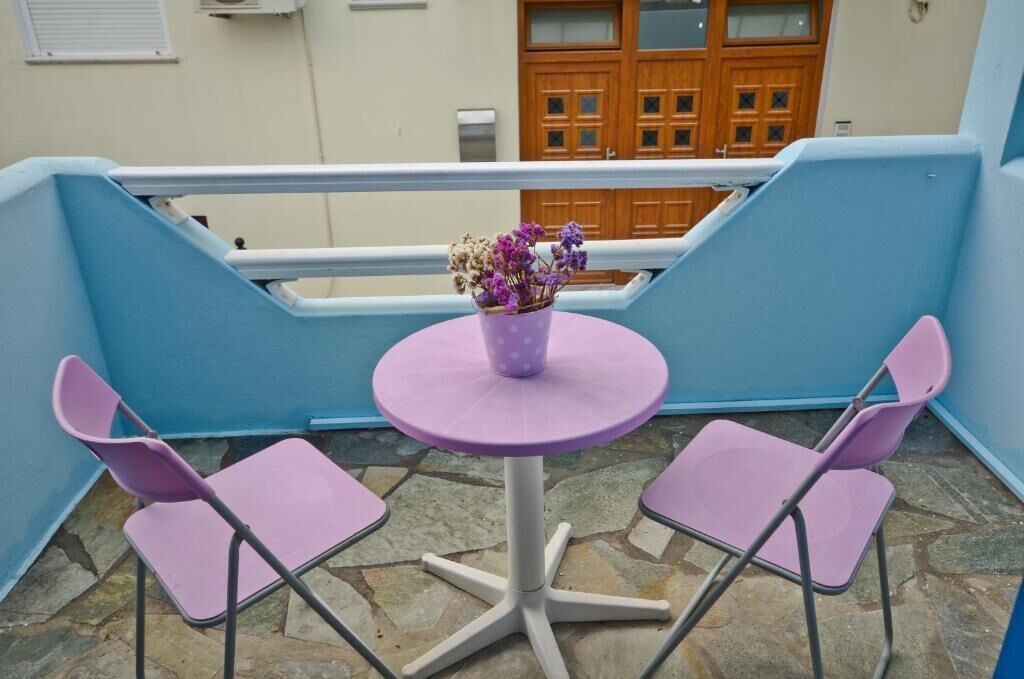 A balcony with two purple chairs and a small round table, a potted plant with purple flowers, and a wooden door in the background.