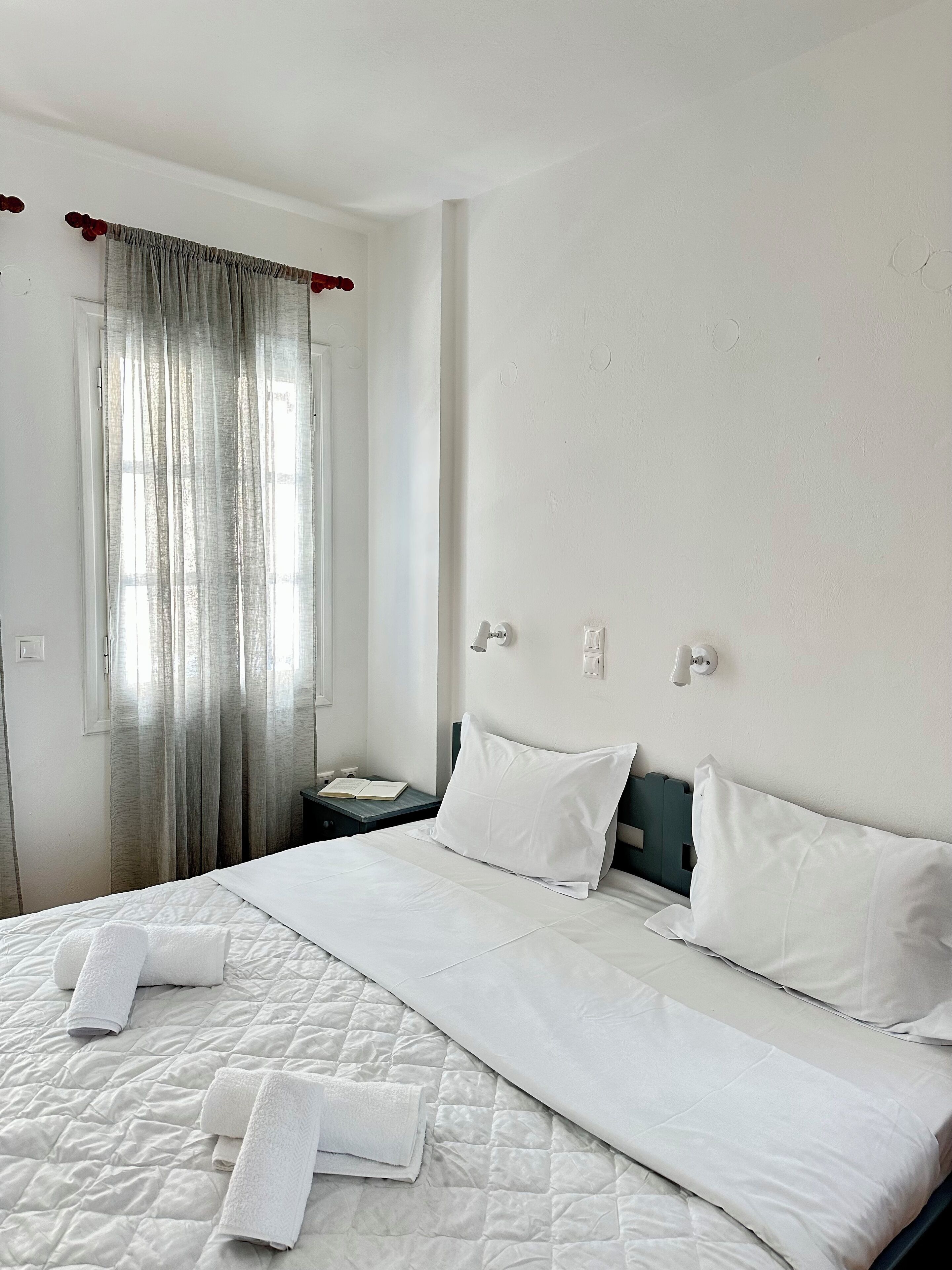 A neatly made bed with white linens and towels, a bedside table with a book, and a window with curtains.